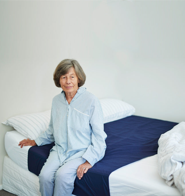 Woman sitting on the edge of a bed which has a Navy bed pad with tuck in wings fitted tucked in above the mattress